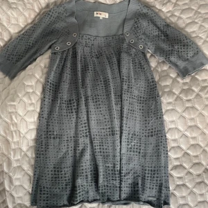 Grå mysig tunika från Billabong - Snygg och mysig, grå tunika från Billabong, med prickigt mönster och trekvartsärmar. Tunikan har en fyrkantig ringning med dekorativa knappar framtill och ett mjukt, skönt material som faller fint. Perfekt för dig som gillar snygga men bekväma plagg. Inköpt för flera år sedan, och flitigt använd, men ser fortfarande bra ut på, förutom dom skavankerna som är specificerade på bilderna. Hålet syns inte när man bör plagget, och nopprorna går att få bort, har själv bara inte rätt verktyg. 