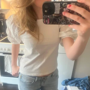 Vit blus med puffärm - Säljer en vit blus med rund hals och korta puffärmar. Toppen är croppad och har en clean, enkel design utan mönster eller detaljer. Materialet känns som bomull och ger en fräsch och lätt känsla. Perfekt för dig som gillar stilrena och trendiga plagg.
