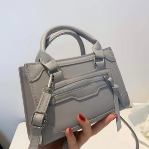 Ljusgrå axelväska med dragkedja - Snygg ljusgrå axelväska i stilren design med både handtag och justerbar axelrem. Väskan har en dekorativ dragkedja framtill och är tillverkad i slätt syntetmaterial. Perfekt storlek för det viktigaste och passar till många outfits.