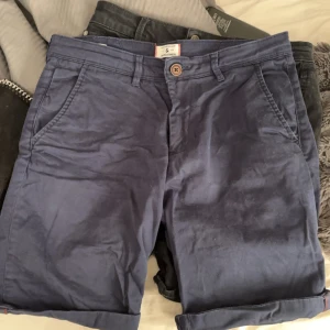 Blåa shorts från Jack & Jones - Snygga marinblå shorts från Jack & Jones med klassisk design, uppvikta benslut och flera fickor fram och bak. Tillverkade i mjuk bomull med knapp och dragkedja i midjan. Perfekta för varma dagar och enkel att matcha med t-shirt eller skjorta.