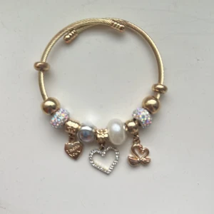 Guldigt armband med berlocker och pärlor - Charmigt armband i guldton med flera berlocker, bland annat hjärtan, glittriga pärlor och en söt rosett. Armbandet har detaljer i vitt och silver samt gnistrande stenar som ger extra bling. Perfekt accessoar för att lyfta din outfit.