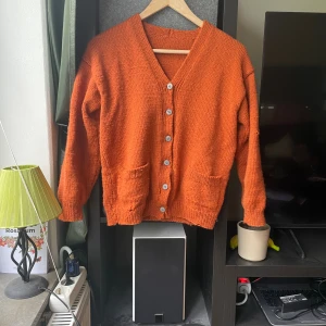 Orange stickad kofta med knappar - Mysig orange stickad kofta med v-ringning och stora silvriga knappar framtill. Koftan har två fickor och ribbade muddar vid ärmslut och nederkant. Har hål och sönder på olika ställen men annars fräscht