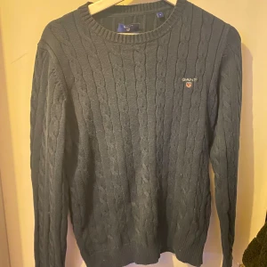 Mörkblå kabelstickad tröja från Gant - Kabelstickad mörkblå tröja från Gant med rund halsringning och broderad logga på bröstet. Tröjan har långa ärmar och ribbade muddar vid ärmslut och nederkant. Perfekt för dig som gillar klassisk och stilren look.