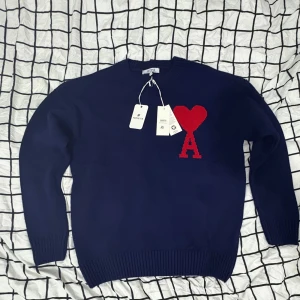 Mörkblå stickad tröja från Ami Paris - Säljer en mörkblå stickad tröja från Ami Paris med röd hjärt- och A-detalj på bröstet. Tröjan har rund hals, långa ärmar och ribbade muddar. Perfekt för dig som vill ha en stilren look. (Rep)