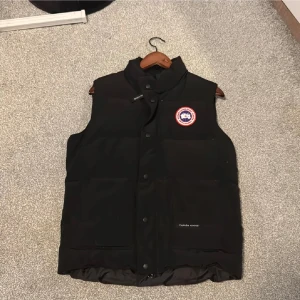 Svart dunväst från Canada Goose - Svart dunväst från Canada Goose med klassisk logga på bröstet. Västen har hög krage, knäppning med både dragkedja och tryckknappar samt två stora fickor framtill. Perfekt för lager-på-lager och riktigt snygg till streetwear.
