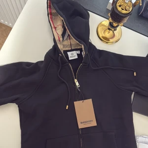 Svart hoodie från Burberry med dragkedja - Svart hoodie från Burberry med klassisk beige rutigt foder i huvan, guldfärgade detaljer och dragkedja framtill. Perfekt för dig som vill ha en stilren och lyxig streetlook.