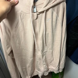 Ljusrosa hoodie från Gymshark - Säljer en ljusrosa hoodie från Gymshark med dragkedja framtill, huva med snörning och två fickor. Tröjan har en stilren look med diskret logga på bröstet och är tillverkad i mjukt bomullsmaterial. Perfekt för chill dagar eller till gymmet.