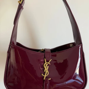 Vinröd axelväska från Saint Laurent - Snygg vinröd axelväska från Saint Laurent i glansigt skinn med ikoniskt YSL-spänne i guld framtill. Väskan har en stilren, kompakt form och bred axelrem som ger en cool vibe. Perfekt för dig som vill ha en statementväska med lyxig känsla.