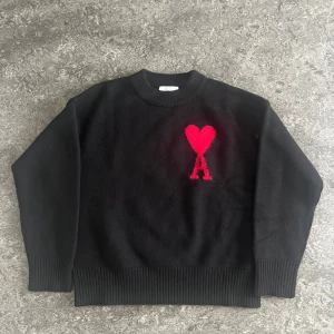 Svart stickad tröja från Ami Paris - Svart stickad tröja från Ami Paris med rund halsringning och ribbade muddar. På bröstet finns ett rött hjärta med bokstaven A, vilket ger en snygg kontrast. Perfekt för dig som gillar stilrena plagg med en twist.