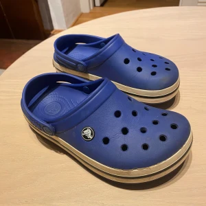 Blå Crocs sandaler med vit kant (barn) - Blå Crocs sandaler med klassisk design, ventilerande hål och vit rand runt sulan. Gjorda i lätt syntetmaterial med justerbar hälrem och Crocs-logga bak. Perfekta för chill dagar eller när du vill ha något bekvämt på fötterna. Storlek 22 centimeter från häl till stortå.