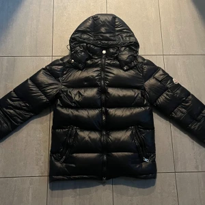 Svart Moncler pufferjacka med huva - Säljer en svart pufferjacka från Moncler med huva och dragkedja framtill. Jackan har två snedställda fickor med dragkedja och Moncler-logga på ärmen. Den är vadderad och har en glansig finish, perfekt för kalla dagar.