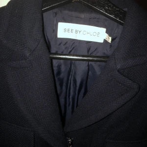 Svart kappa från See by Chloé - Snygg svart kappa från See by Chloé med klassisk krage, fyra fickor framtill och diskret fiskbensmönster i tyget. Jackan har dragkedja och är helfodrad, perfekt för dig som vill ha en stilren och tidlös look.