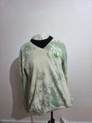 Grönvit v-ringad tröja G.C. Spaarnwoude - Säljer en v-ringad långärmad tröja från G.C. Spaarnwoude i storlek M. Tröjan har ett grönt och vitt batikmönster och en broderad logga på bröstet. Ribbstickade muddar vid ärmslut och nederkant. Perfekt för dig som gillar sportig och chill stil.