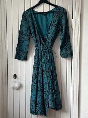 Turkos omlottklänning från Noa Noa - Härlig omlottklänning från Noa i perfekt färgkombo - mörk turkos och mörkbrunt. Klänningen har broderade trekvartsärmar och tunt knytband i midjan som kan knytas loose i ryggen eller dubbelt varv fram i midjan. Materialet är väldigt följsamt i viskos crêpe. Midi längd. Passar även S och M. . 