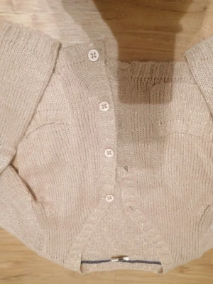 Glittrig beige kofta från Pomp de Lux - Säljer en beige glittrig kofta från Pomp de Lux i storlek 134/140. Koftan är stickad med ribbade muddar och har vita knappar framtill. Den har en mjuk och glansig finish som ger extra stilpoäng. Perfekt för dig som gillar lite extra glitter i vardagen. Passar även dig som bär XXS/XS. 😊