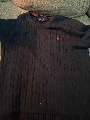  Äkta Svart stickad tröja från Polo Ralph Lauren - Svart stickad tröja från Polo Ralph Lauren med klassiskt kabelstickat mönster och röd broderad logga på bröstet. Rund halsringning och ribbade muddar vid ärmslut och nederkant. Perfekt för dig som gillar stilrena och tidlösa plagg.