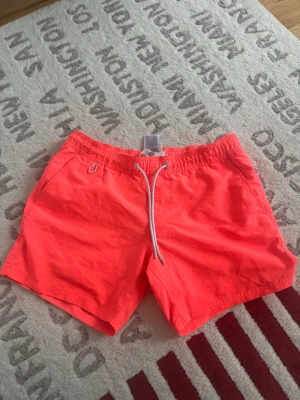 Neonorange badshorts från FSBN Beach - Säljer ett par neonorange badshorts från FSBN Beach i storlek XS. Shortsen har elastisk midja med vit snörning, innerbyxa i mesh och en bakficka med dräneringshål. Perfekta för strandhäng och poolparty!
