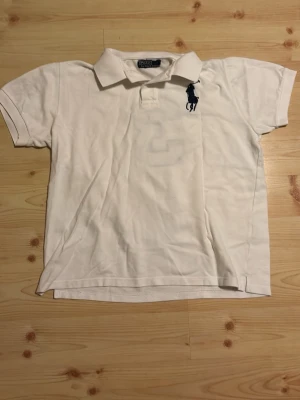 Vit polotopp från Polo Ralph Lauren - Säljer en klassisk vit polotopp från Polo Ralph Lauren i storlek M. Toppen har korta ärmar, krage och knappar framtill. På bröstet finns den ikoniska Polo-loggan i mörkblått och en stor mörkblå siffra 3 på ryggen och ärmen. Perfekt till en chill och sportig stil. Denna är lite äldre version också som jag fick av min morfar så den är lite äldre men i perfekt skick
