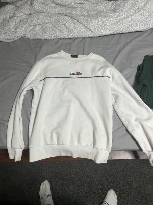 Vit sweatshirt från Ellesse, M - Säljer en vit sweatshirt från Ellesse i storlek M. Tröjan har rund hals, svart linje över bröstet och Ellesse-logga broderad framtill. Tillverkad i mjuk bomull och har långa ärmar med ribbade muddar. Perfekt för en clean och sportig look. Storlek M men passar S också