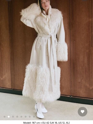 Vit lång kappa med pälsdetaljer - Lång vit kappa med fluffiga pälsdetaljer på krage, ärmslut och nederkant. Kappan har bälte i midjan, dubbelknäppning och stora slag. Perfekt statement-plagg för dig som vill sticka ut med en lyxig och trendig look. Jackan är helt oanvänd och nyköpt. Köpt för 1499kr