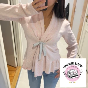 Ljusrosa omlottkofta med rosett - Supersöt ljusrosa omlottkofta från Unique Shop med volangkant och grå knytrosett framtill. Koftan har lång ärm och är tillverkad i mjuk bomullsmix som känns skön mot huden. Perfekt att styla med jeans för en chill look.