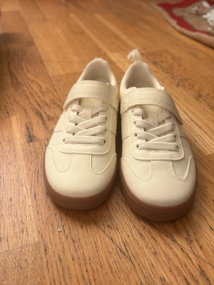 Beige sneakers med bruna sulor - Snygga beige sneakers med bruna platta sulor och rund tå. Skorna har både snörning och kardborreband för extra smidig passform. De är nästan helt nya och är perfekt present för barnen nu inför jul 