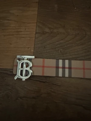 Rutig Burberry bälte - Säljer ett snyggt Burberry bälte med klassiskt rutig design i beige, rött, svart och vitt. Bältet har ett metallspänne med TB-logga i silverfärg. Materialet är skinn med textil på ovansidan. Perfekt för att lyfta din outfit med en ikonisk detalj.