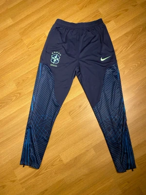 Nike Brasil träningsbyxor blå - Snygga blå träningsbyxor från Nike med Brasil-logga och mönstrade detaljer längs benen. Byxorna har elastisk midja och en fräsch design med ljusgrön Nike-swoosh. Perfekta för dig som gillar fotboll och vill ha något sportigt och bekvämt.