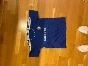 Chelsea fotbollströja Torres #9 - Blå Chelsea fotbollströja med vita detaljer, tryck med Samsung på framsidan och Torres #9 på ryggen. Klassisk design med klubbmärke på bröstet och korta ärmar. Perfekt för dig som älskar fotboll och vill visa ditt stöd för Chelsea.