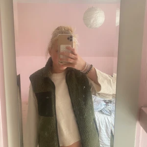 väst  - Mörkgrön fleeceväst från Sail Racing med svart bröstficka med dragkedja och svarta detaljer vid fickor och krage. Västen har en mysig teddylook och är perfekt att slänga över en hoodie eller tröja för extra värme.