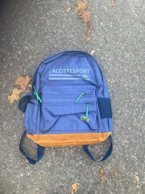 Blå ryggsäck från Lacoste Sport - Säljer en snygg blå ryggsäck från Lacoste Sport med bruna mockadetaljer i botten och gröna dragkedjor. Väskan har flera fack, meshficka på sidan och justerbara axelremmar. Perfekt för skola eller utflykter, med coolt Lacoste-tryck framtill.