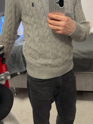 Grå stickad half zip tröja Polo Ralph Lauren - Säljer en grå stickad tröja från Polo Ralph Lauren med klassiskt kabelmönster och half zip-dragkedja. Tröjan har långärmade ribbade muddar och en broderad logga på bröstet. Perfekt för dig som gillar stilren och tidlös design. Jag är 180 och väger 74 kilo 