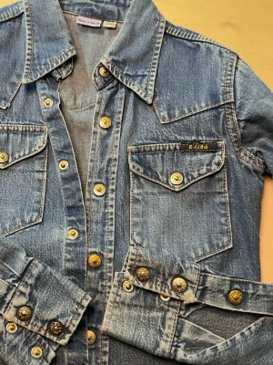 Blå jeansjacka från Gul & Blå - Säljer en klassisk blå jeansjacka från Gul & Blå med snygga guldiga knappar och två bröstfickor med lock. Jackan har markerad krage, kontrastsömmar och cool vintagekänsla. Perfekt för dig som gillar retrostil och denim.