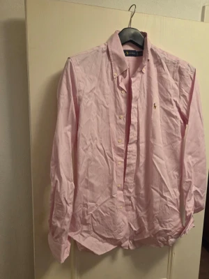 Rosa skjorta från Ralph Lauren - Säljer en klassisk rosa skjorta från Ralph Lauren med button-down krage och broderad logga på bröstet. Skjortan har långa ärmar och knappar framtill. Materialet är mjuk bomull och passformen är normal. Perfekt för dig som gillar stilrena plagg med ikonisk design.