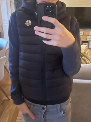 Svart dunväst från Moncler - Säljer en svart dunväst från Moncler med klassisk logotyp på bröstet. Västen har quiltad design och dragkedja framtill. Perfekt att slänga över en hoodie för en chill och snygg look. Passar dig som gillar stilrena och funktionella plagg.