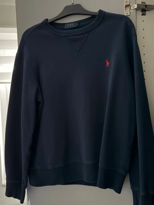 Mörkblå sweatshirt från Polo Ralph Lauren - Snygg mörkblå sweatshirt från Polo Ralph Lauren med klassisk röd logobrodyr på bröstet. Tröjan har rund hals och långa ärmar, perfekt för chill dagar. Mjuk bomullskvalitet och normal passform. Enkelt stilren och passar till jeans eller joggers.