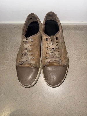 Bruna sneakers från Lanvin - Säljer ett par bruna sneakers från Lanvin med rund tå och platt sula. Skorna har snörning och är tillverkade i mocka med detaljer i skinn på tån. Snygg och enkel design med beige snören och svart innersula. Perfekt för dig som gillar stilrena sneakers.