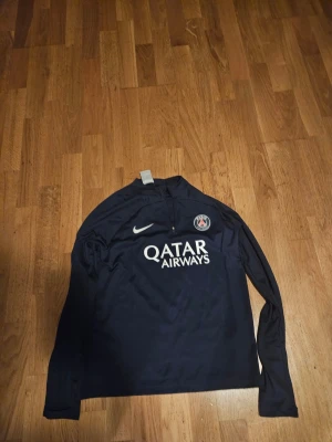 PSG Nike Dri-Fit träningströja s - Mörkblå långärmad träningströja från Nike med PSG-logga och Qatar Airways-tryck på bröstet. Tröjan har Dri-Fit material som andas och en liten half zip vid halsen. Perfekt för dig som gillar fotboll och vill ha något snyggt på träningen.