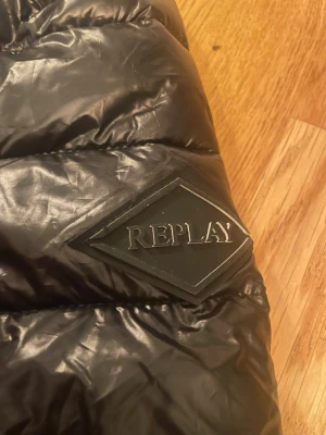 Svart pufferjacka från Replay - Säljer en svart pufferjacka från Replay med huva och dragkedja framtill. Jackan har ett glansigt yttermaterial och Replay-logga på ärmen. Perfekt för kalla dagar och har en modern, sportig look.