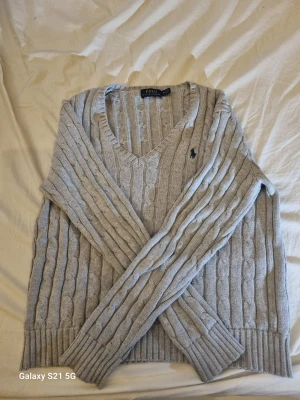 Kabel stickad Polo Ralph Lauren tröja - Säljer en grå kabelstickad tröja från Polo Ralph Lauren i storlek M. Tröjan har v-ringad hals, långärmade ribbade muddar och ett broderat mörkblått logomärke på bröstet. Perfekt för dig som gillar klassisk och stilren design.