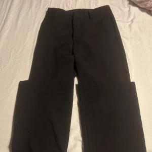 Svarta bootcut kostymbyxor - Snygga svarta kostymbyxor med bootcut-snitt och klassiska bälteshällor. Byxorna har en stilren och enkel design som passar till många olika looks. Materialet känns mjukt och har en lätt stretch för extra komfort. Perfekta för dig som gillar en tidlös och clean stil.