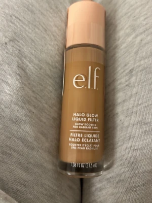e.l.f. Halo Glow Liquid Filter Medium - Glow booster i flytande form från e.l.f. i färgen Shade 4 Medium. Kommer i en genomskinlig flaska med persikofärgat lock och innehåller 31,5 ml. Ger huden en strålande finish och kan användas som bas eller highlighter.Den är inte öppnad