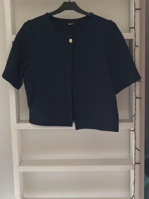 Mörkblå kort kofta från gina tricot - Kort och stilren mörkblå kofta från gina tricot med rund hals och en dekorativ guldfärgad knapp framtill. Koftan har korta ärmar och en rak passform som ger en clean look. Perfekt att slänga över en topp för en enkel och snygg outfit.