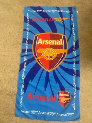 Arsenal badhandduk i blått - Säljer en stor blå badhandduk med Arsenal-logga och texten 'Arsenal' i rött och gult. Handduken har ett coolt mönster med kanon och klubbens emblem i mitten och längs kanterna. Perfekt för dig som älskar fotboll och vill visa ditt lag på stranden.