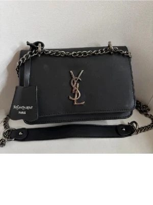 Svart handväska - Säljer en stilren svart handväska med silverfärgad kedja och ikonisk YSL-logga framtill. Väskan är tillverkad i skinn och har vinrött innerfoder med flera fack för smart organisering. Perfekt storlek för det viktigaste och en snygg detalj till din outfit.