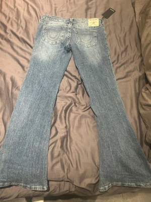Blå bootcut jeans från True Religion - Säljer ett par blå jeans från True Religion med klassiska fickor och tydliga sömmar bak. Modellen har bootcut-ben och låg midja, vilket ger en snygg siluett. Jeansen är tillverkade i denim och har en tvättad look med ljusare partier framtill. Köptes för 1399 kr, vid snabb affär kan jag tänka mig att sänka pris! Prislapp kvar!
