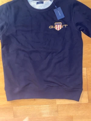 Mörkblå sweatshirt från GANT - Snygg mörkblå sweatshirt från GANT med broderad logga och sköld på bröstet. Tröjan har rund hals och ribbade muddar vid ärmslut och nederkant. Tillverkad i mjuk bomull som är skön att ha på sig. Perfekt för dig som gillar klassisk stil med en sportig touch.tröjan är sprillans den är inte använd en gång.