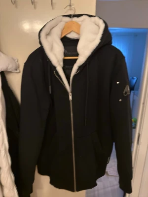 Svart pälsfodrad jacka från Moose Knuckles - Säljer en svart jacka från Moose Knuckles med stor huva fodrad i mjuk vit fuskpäls. Jackan har dragkedja framtill, fickor och en snygg logga på ärmen. Perfekt för kalla dagar när du vill vara både varm och stilren och såklart helt äkta.