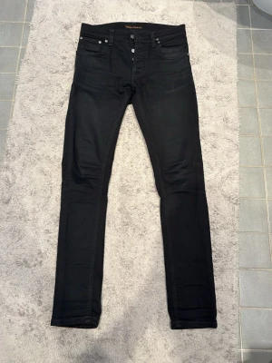 Svarta Nudie Jeans Tilted Tor W28 L30 - Säljer ett par svarta Nudie Jeans Tilted Tor i modellen Dry Ever Black. De har klassisk femficksdesign, skinny passform och är tillverkade av 99% ekologisk bomull och 1% elastan för extra stretch. Perfekta om du gillar stilrena och tajta jeans.