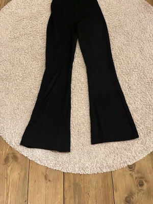 Svarta bootcut-byxor från Gina Tricot - Svarta bootcut-byxor från Gina Tricot i storlek S. Byxorna har en hög midja och är tillverkade i ett mjukt och stretchigt syntetmaterial som sitter skönt. Perfekt för dig som gillar en klassisk och stilren look med lite extra vidd nedtill.